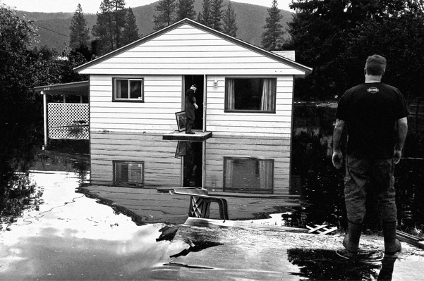 Person standing on floating board looking at a flooded house.