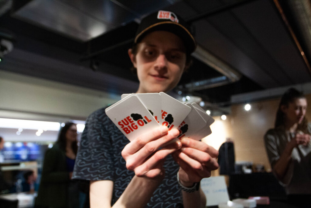 A person holds a hand full of cards with Sue Big Oil logo on the back
