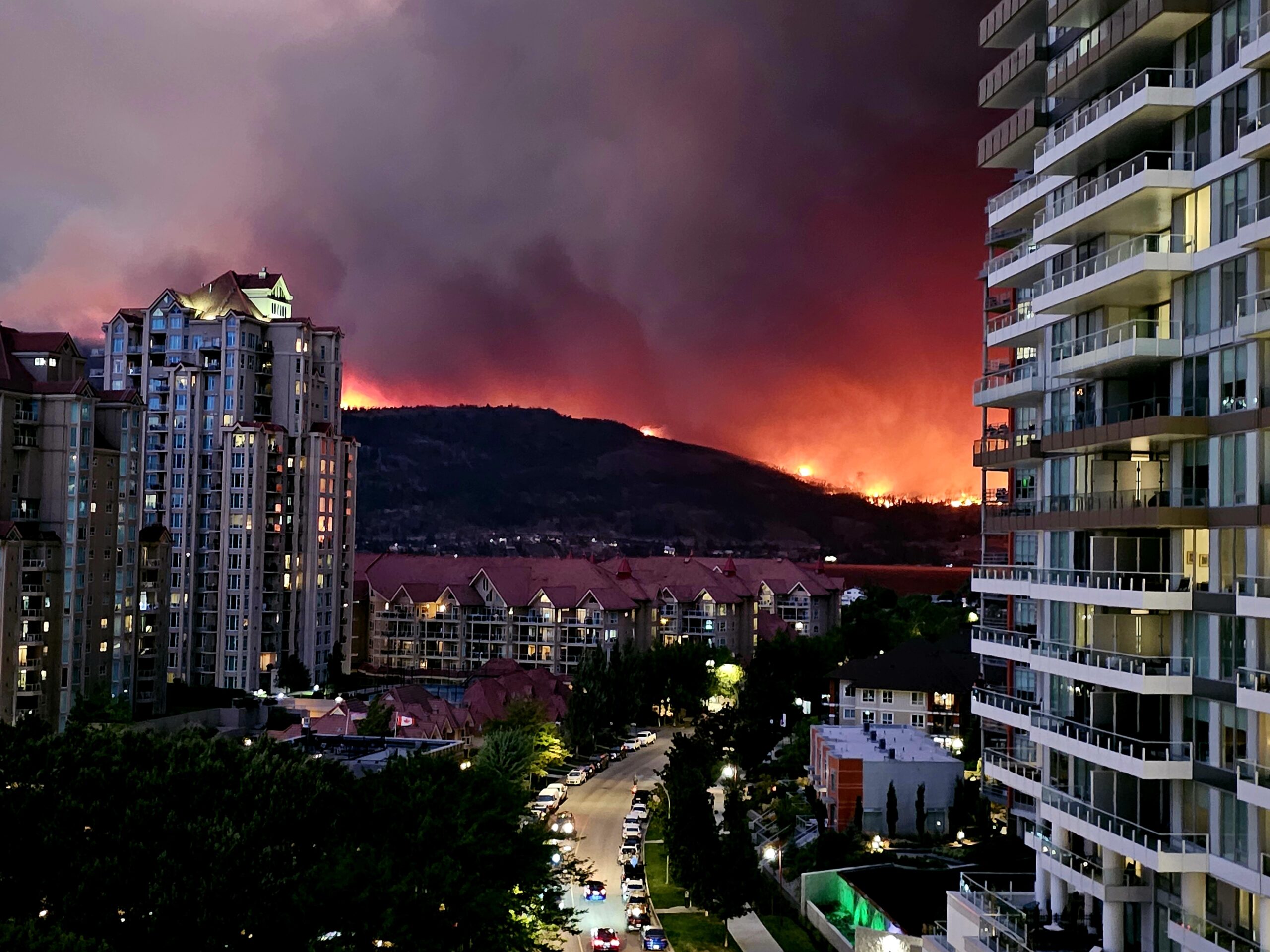 Wildfire consuming a hillside just outside Kelowna, BC with urban buildings in foreground.