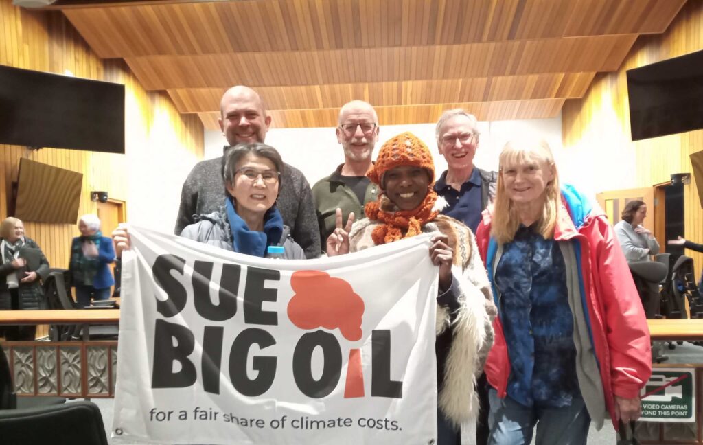 A group of 6 people gathered in Saanich Council offices, holding a "Sue Big Oil" sign