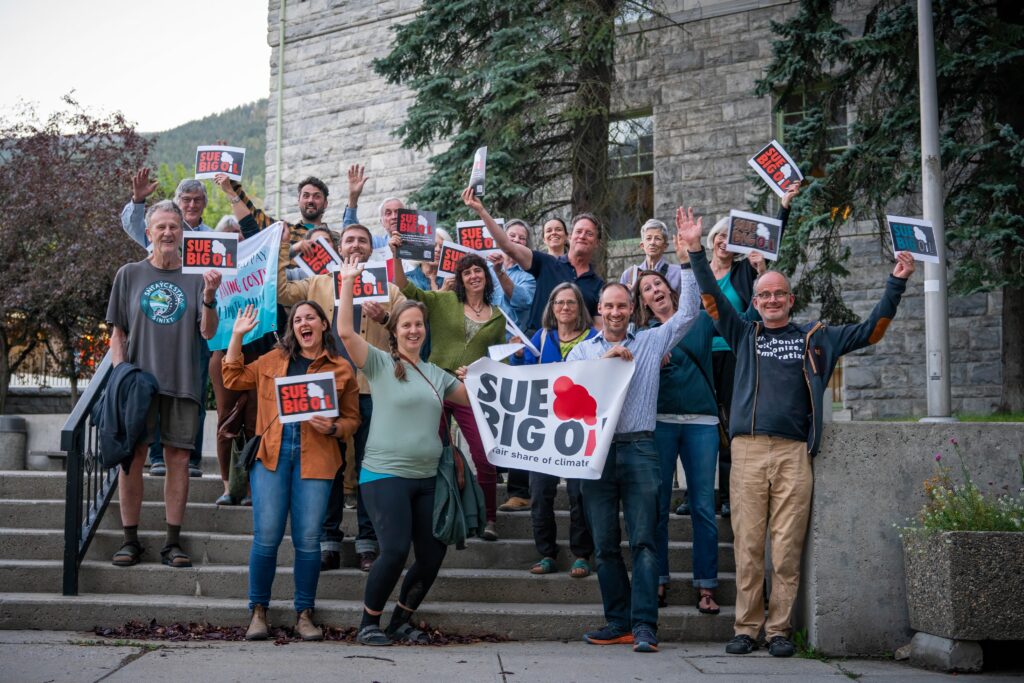 A group of 15-20 supporters holding Sue Big Oil banners on the steps of Nelson City Hall