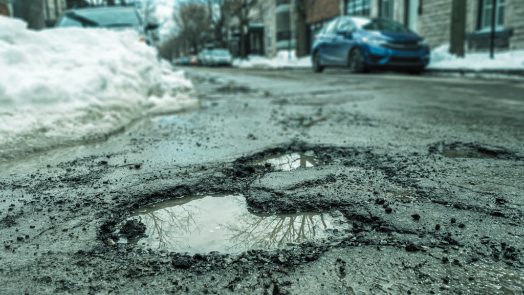 Pothole in an urban street with snow piles and parked cars in the background