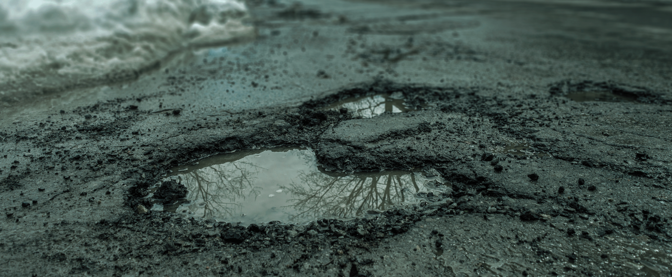 A pothole on an urban street with snow piled next to parked cars in the background